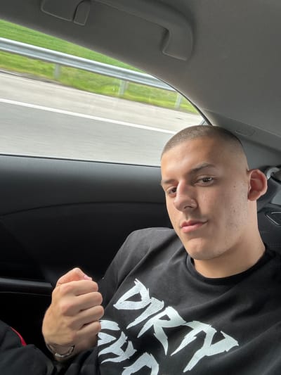 A man with short-cropped hair is in the passenger seat of a car, posing for a photo. He is wearing a dark-colored long-sleeved shirt with a white graphic print. His right hand is clenched in a fist. A car window shows passing scenery consisting of a road and greenery. The location in the city of Svoboda, Bulgaria, is not identifiable based on the visual content of the image.