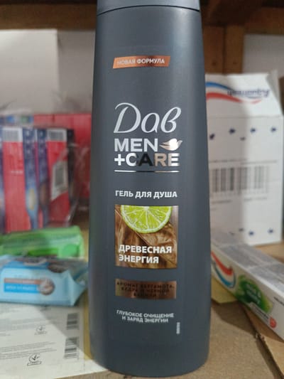 A Dove Men + Care shower gel bottle is prominently displayed in the center of the frame. The bottle is dark gray with silver and white lettering. The top of the bottle has an orange label that says "НОВАЯ ФОРМУЛА" (New Formula). Below the brand name, it reads "MEN + CARE". Further down, in smaller text, it says "ГЕЛЬ ДЛЯ ДУША" (Shower Gel). A circular image of a lime slice is overlaid on a wooden background, beneath which are the words "ДРЕВЕСНАЯ ЭНЕРГИЯ" (Wood Energy). Below this, smaller text reads "АРОМАТ БЕРГАМОТА, КЕДРА И ЧЕРНОЙ СПИЧИ" (Scent of Bergamot, Cedar and Black Pepper). At the bottom of the bottle, it states "ГЛУБОКОЕ ОЧИЩЕНИЕ И ЗАРЯД ЭНЕРГИИ" (Deep Cleansing and Energy Boost). A small serial number "90807810" is visible on the lower right side of the bottle.
The background is blurred but suggests a retail or storage environment. To the left of the Dove bottle, partially visible, are packages of what appear to be wet wipes and other hygiene products. To the right, a partially visible toothpaste box with red, white, and blue stripes is present. The overall lighting is moderate.
The image does not provide any information that would allow for its localization within the city of Berdiansk, Ukraine.
