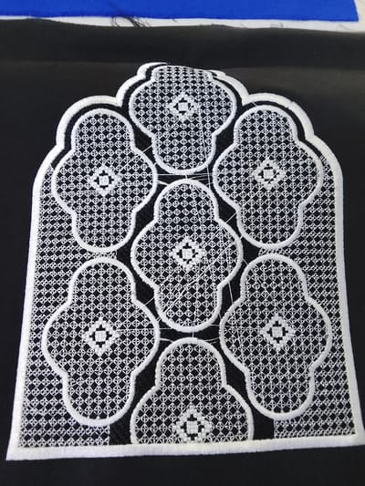 The image shows a close-up of white lace embroidery on a black fabric. The embroidery is in the shape of an arched window with seven clover-like shapes within it, each containing a smaller diamond-shaped design. The embroidery has a thick, outlined border. The fabric is laid flat on a surface, with a hint of blue fabric and white material visible at the top edge of the frame, suggesting it might be part of a larger textile or garment. The lighting is bright, casting some faint shadows. The setting appears to be indoors, possibly a workspace or a home where sewing or embroidery is being done. There is no discernible text in the image. The focus is entirely on the intricate detail of the lace embroidery and its contrast against the dark background.