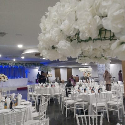 The image displays the interior of a large event hall, configured for a formal gathering in Birmingham, United Kingdom; the specific venue within the city is not identifiable. The scene is dominated by a white aesthetic, featuring numerous round tables covered with white tablecloths and surrounded by white Chiavari-style chairs. Each table is set with white plates, cutlery, napkins, and bottles of various beverages. In the upper foreground, a large, spherical artificial floral arrangement composed of dense white blossoms attached to a green frame partially obstructs the view. In the background, a stage or elevated platform is visible, adorned with blue drapes and a long white floral garland along its upper edge, illuminated by blue accent lighting. Several other large, artificial white floral trees or arrangements are positioned throughout the hall. The floor is dark and reflective. Approximately six individuals are visible within the space. One person in dark clothing stands in the mid-ground, apparently engaged with a mobile phone. Another individual, dressed in a reddish-brown garment, stands to the right of the frame. No specific actions or direct interactions are clearly discernible among the present individuals.