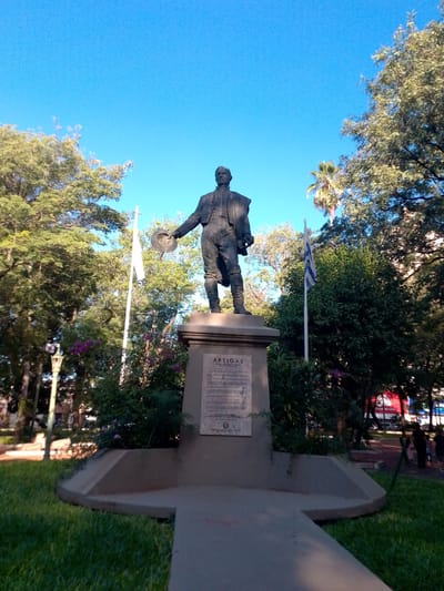 This is an outdoor scene, under a bright blue sky. The focal point is a statue of José Gervasio Artigas, a historical figure, standing on a stone pedestal. The pedestal is inscribed with text and is surrounded by greenery, including flowers and bushes. Two flags are visible to either side of the statue: one is plain white, and the other features blue and white stripes, resembling the flag of Uruguay. Trees provide a backdrop, with a palm tree also visible in the distance. The setting appears to be a park in the city of San Lorenzo, Paraguay. In the background, hints of a city street can be seen. There are some pedestrians in the distance.