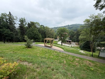 The image depicts an outdoor public space in Świeradów-Zdrój, Poland, on a cloudy day. The foreground features a grassy slope with a path winding through it. A wooden gazebo-like structure with benches is positioned on the path, and a solitary bench is also visible nearby. Further along the path, a broader paved area with tiered seating leads towards buildings and trees in the middle ground. In the background, rolling hills covered in dense green forest stretch towards the horizon under a gray sky. Several indistinct figures are walking on the paths, and trees are abundant throughout the scene, indicating a park or recreational area within the town.