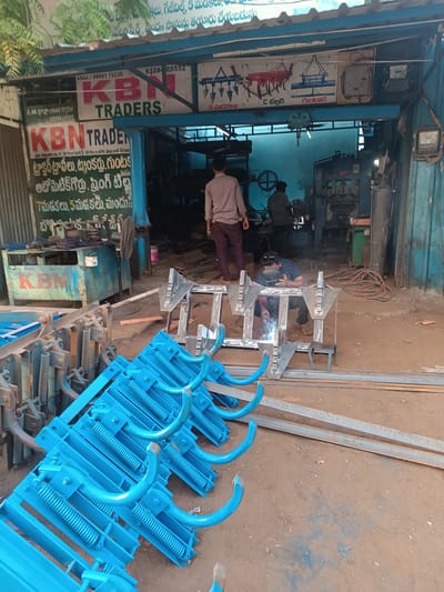 This image depicts an active metal fabrication workshop identified as "KBN TRADERS" in Anantapur, India. The location is confirmed by Telugu script on the prominent signboard, which also specifies agricultural equipment like "Tractor Tools," "C Tiller," and "Guntaka."

In the foreground, a large quantity of newly manufactured or refurbished agricultural components, painted bright blue with springs and curved tines, are stacked on the dirt ground. Directly behind these parts, in the midground, a man wearing a dark blue patterned shirt and sandals is actively welding a raw metal frame. He is crouched, wearing a welding mask, with visible sparks emanating from the welding process. To his right, coiled welding cables are present.

Further into the open-front workshop, two additional men are visible. One, wearing a light brown shirt and dark pants, stands partially obscured, facing away from the viewer. The other, dressed in a dark grey shirt, is operating heavy machinery, possibly a press or shear, in the background. The workshop interior contains various industrial tools and equipment, including a light blue workbench, an industrial fan, metal stock, and a tall gas cylinder. An electrical meter box and associated wiring are mounted on the exterior right wall. The floor throughout the scene is unpaved dirt. Above the workshop entrance, signboards in English and Telugu display the "KBN TRADERS" name, contact numbers (98661 75236, 83284 05552), and illustrations of agricultural implements. The overall environment is a bustling, functional industrial space.
