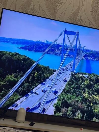 The image is an aerial view of a large suspension bridge, likely the Bosphorus Bridge in Istanbul, Turkey, spanning a wide body of blue water. The bridge is crowded with traffic, including cars, trucks, and buses. Lush green trees and forested hillsides flank the approach to the bridge on both sides. In the distance, a city skyline with numerous buildings is visible. The sky is a clear blue. The photograph is taken from a high angle, giving a comprehensive view of the bridge and its surroundings. There is no indication of the city of Marchevo, Bulgaria in the image.