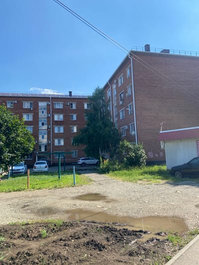 The image shows a brick apartment building with multiple floors and windows. There are several cars parked in front of the building, including white sedans and a darker colored vehicle. A gravel path runs alongside the building, with a large puddle of water on it, indicating recent rain. There is also an area of dark soil with some green shoots, suggesting a garden or undeveloped land. Trees and green grass are visible around the building and path. The sky is clear and blue, with a few wispy clouds. The sun appears to be shining, casting shadows and illuminating the scene. The time of day appears to be daytime. There is no visible text in the image.