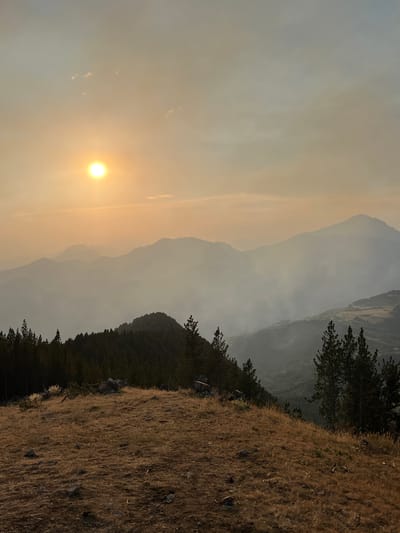 The image depicts a panoramic view of a mountainous landscape at sunset, likely in the vicinity of San Juan de Plan, Spain, a town known for its natural beauty and proximity to the Pyrenees. The scene is characterized by a hazy, golden sky as the sun, a bright orb, hangs low on the horizon, casting a warm glow over the environment.

In the foreground, a dry, grassy hill slopes downwards, dotted with scattered rocks. Several pine trees, some with dark green needles, stand on this ridge, their silhouettes stark against the lighter background. The mid-ground reveals a dense forest of pine trees clinging to a steep incline, their dark forms creating a strong contrast with the softer hues of the distant mountains.

Beyond the immediate forest, layers of mountains recede into the distance, their forms softened by a visible haze or smoke that lends a dreamlike quality to the scene. This atmospheric effect obscures finer details of the landscape, presenting a gradient of muted blues and grays. In the valleys between the mountains, hints of human settlement or agricultural land can be faintly discerned, suggesting the presence of scattered structures or cultivated fields. The overall impression is one of serene, wild beauty, possibly influenced by natural phenomena such as forest smoke or atmospheric conditions typical of mountainous regions.