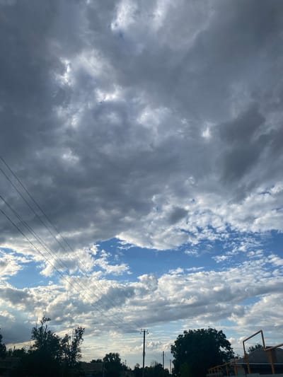 The media file is a photograph of a dynamic sky with varying cloud formations. The majority of the frame is filled with large, dark, and ominous storm clouds on the left, interspersed with patches of bright white and light grey cumulus clouds. To the right, the sky transitions to a clear blue, broken by streaks of smaller, wispy clouds that catch the sunlight, creating a dramatic contrast.
Visible in the lower right portion of the image are silhouetted trees, suggesting an outdoor setting with natural elements. A power line with multiple wires stretches diagonally across the frame, disappearing into the clouds and trees, hinting at a semi-urban or rural environment. A hint of a building structure with a yellow railing is visible at the top right edge, providing a further clue to the location.
The lighting suggests it is either early morning or late afternoon, with the sun's rays filtering through the clouds, creating shafts of light and casting shadows. The overall mood conveyed is one of dramatic weather, with the juxtaposition of dark storm clouds and bright, sunlit skies. There is no visible text in the image. Given the provided location context of Taganrog, Russia, the scene likely depicts the sky over that region.