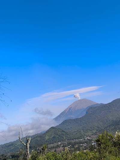 This image captures a dramatic volcanic eruption near Ranupane, Indonesia. A tall, conical volcano, likely Mount Semeru, is prominently featured in the background, emitting a plume of ash and smoke that billows into the bright blue sky. The volcano's slopes are covered in a layer of gray ash, contrasting with the lush green vegetation of the surrounding mountainsides. In the foreground, a rural landscape unfolds, with scattered houses and farms nestled among rolling hills. The scene is dominated by the raw power of nature, with the erupting volcano serving as a stark reminder of the earth's geological forces. The clear sky and sunlight suggest a daytime setting, though the smoke plume partially obscures the upper reaches of the volcano.