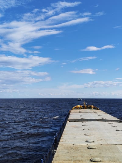 A wide-angle, eye-level view captures a cargo ship sailing on a deep blue, slightly choppy body of water under a bright blue sky with scattered white clouds. The ship's deck, constructed of weathered, light gray metal, occupies the foreground and extends into the distance, punctuated by rows of circular hatches with visible handles. The ship's stern, painted in yellow and blue, is visible at the far end of the deck. In the distance, two smaller vessels are visible on the horizon, appearing as dark shapes against the water. The scene is set on a waterway, possibly a sea or large river, with no visible landmasses or specific city landmarks that would indicate the location in Dor, Russia. The overall impression is one of open water travel under fair weather conditions.