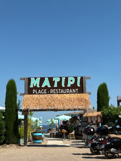 The image shows the entrance to a beachside establishment named "MATIPI PLAGE • RESTAURANT" in Théoule-sur-Mer, France. The sign, with large teal lettering on a dark brown background, is framed by a thatched roof structure. To the left and right of the entrance are tall, dark green cypress trees. In front of the establishment, two black scooters are parked. Behind the entrance, beach umbrellas, tables, and chairs are visible, suggesting an outdoor dining or seating area. People are seated at tables further back, closer to the sea. The ground is a mix of sand and gravel. The sky is clear and blue.