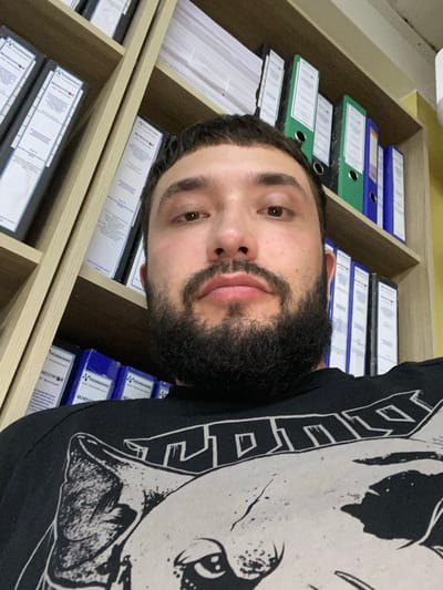 A close-up, low-angle, selfie-style photograph captures a man with a beard, positioned in front of bookshelves filled with binders. The man is wearing a black t-shirt with a graphic print. The environment appears to be an office or study, with the bookshelves forming the backdrop. The man's face, partially obscured by his beard, is the central focus. The binders on the shelves are of various colors, including white, blue, and green, and some have labels with Russian text. The lighting suggests an indoor setting.