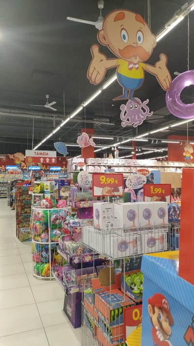 The image depicts the interior of a toy store, likely a section of a larger supermarket, located in Pylaia Municipal Unit, Greece, as suggested by the presence of Greek text on signage. In the foreground, shelves are filled with various toys, including stuffed animals, board games, and smaller packaged items like fans. Prominently displayed are several pricing signs, with one reading "9,99 €" and another "1,99 €". Hanging from the ceiling are decorative elements, including cartoon figures and inflatable rings, which are common in children's retail environments. The store is well-lit with overhead fluorescent lighting and ceiling fans are visible. The overall impression is a brightly lit, well-stocked retail space designed to attract shoppers, particularly families with children. There are no individuals clearly visible in the frame, but the arrangement of merchandise suggests an active retail environment.