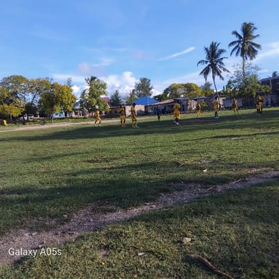 The image shows a group of young men playing soccer in an open field in the village of Nungwi, located on the north-western coast of the island of Unguja, Zanzibar, Tanzania. The coordinates provided are -5.726, 39.301, which correspond to a location near the shore of the Indian Ocean. The scene is captured from a distance, showcasing a wide expanse of green grass. A couple of men are seen dribbling the ball and passing it among themselves. Some of the young men are standing on the field, observing the play, while others are positioned in the sidelines. Behind the men are residential buildings with roofs made of corrugated iron sheets. There are palm trees and bushes in the background. The scene is illuminated by the bright afternoon sunlight, casting long shadows on the field. There are no spectators in the image. The image was taken by a mobile phone, with the phone's brand name "Galaxy A05s" visible in the bottom left corner.