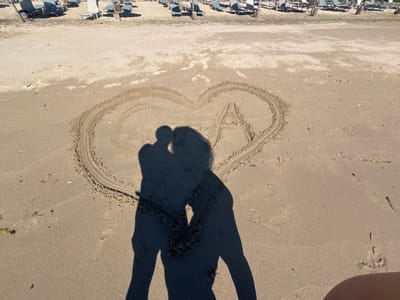 In Rezovo, Bulgaria, on a sunlit beach, a heart shape has been drawn in the sand. Inside the heart, the letters "C" and "A" are visible. The shadows of two people, one appearing to lean in for a kiss, are cast within the heart, obscuring the letters. In the background, rows of unoccupied beach chairs are arranged on the sand, with the sea not visible. The overall scene depicts a romantic moment captured on a beach.
