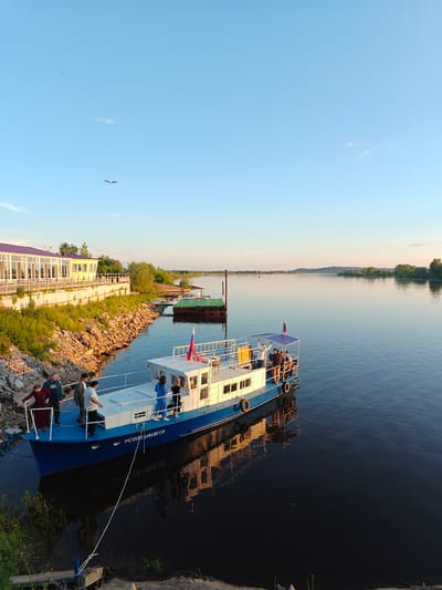 This detailed description of the image is provided from a forensic, matter-of-fact perspective. The image depicts a wide river scene in Nikolo-Beryozovka, Russia, under a clear, light blue sky during what appears to be late afternoon or early evening. The lighting is warm and directional, suggesting golden hour, with the sun illuminating the left riverbank and parts of the boat. In the upper portion of the frame, the sky is a gradient of light blue, slightly lighter towards the horizon. A single, small, distinct white cloud is present in the upper-center, with a few faint wisps of clouds visible elsewhere. A dark bird with spread wings is captured in flight in the upper-left quadrant, with a smaller, barely visible speck in the sky further left. The majority of the lower half of the image is occupied by a wide, calm river with dark blue-green water, reflecting the sky and the warm sunlight, particularly near the foreground. Prominently featured in the mid-ground is a leisure boat with a blue hull and a white cabin, docked close to the left bank. A white rope tethers the boat to a small post on the bank. The registration "УС0231RUS18" is clearly visible in white lettering on the blue hull near the bow. Several black tires serve as fenders along the boat's side. The boat's reflection, including the hull, cabin, and the inverted registration, is distinctly visible in the still water. A Russian flag (white, blue, red horizontal stripes) flies from a pole at the stern of the boat. The upper deck features a blue canopy structure. Approximately eight to ten individuals are visible on and around the main boat. Three people, two men and one woman in a maroon dress or coat, are standing on the boat's main deck near the bow, facing the bank, with the woman holding onto a railing. On the upper deck under the blue canopy, several other people are present, including two women in blue and black attire standing on the starboard side. On the rocky bank directly adjacent to the boat, a man in a dark jacket stands, appearing to interact with those on board. The left riverbank, sloping towards the water, is covered with green vegetation, tall grasses, and exposed rocks, all strongly lit by the warm, golden sunlight. Further down this bank, a light yellow building with numerous large glass windows and a distinctive purple roof