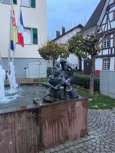 This image depicts a public square in Oppenau, Germany, featuring a functional fountain and a bronze sculpture. The scene is observed under an overcast sky, suggesting a cloudy day or dusk.

In the foreground, a multi-tiered fountain comprises a large, rectangular basin made of reddish-brown stone, exhibiting prominent dark vertical streaks from water runoff. Water actively flows from several jets on the left side of the basin, creating foam and surface agitation. Positioned on a raised platform within the basin, towards the right, is a patinated bronze sculpture group of three individuals. The figures appear to represent traditional laborers or townspeople.

The leftmost figure is a seated or crouching male with a mustache, wearing an apron, and manipulating an object in a small trough or basin. To his immediate right and slightly behind, a female figure stands, her head covered with a cloth. The third figure, a male, stands to the right, wearing a hat and holding what appears to be a small barrel or bucket. He is depicted with one foot resting on a smaller barrel or stump. Text engraved into the fountain's stone base is partially visible, including "SPARKASSE OPPENAU".

To the left of the fountain, a light-colored multi-story building with green-shuttered windows stands, displaying two flags on poles: a yellow flag with a red emblem and German text, and the French national flag (blue, white, red). A third, dark blue flag is partially visible behind the yellow flag. Adjacent to this building, behind the fountain, is a white utility cabinet on a patch of grass.

To the right of the fountain, a traditional half-timbered building with white infill, dark brown timbers, and red window trim is visible, featuring a dark-tiled roof. Several pollarded deciduous trees with sparse green-yellow leaves and bare branches are interspersed between the fountain and the buildings, indicating late autumn or early winter. The ground surface is paved with grey cobblestones, with sporadic weeds growing from the crevices.