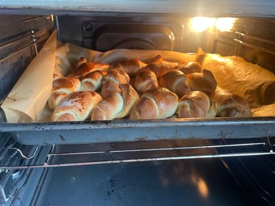 A close-up, inside-oven shot captures a baking tray laden with golden-brown croissants. The croissants are arranged on parchment paper, their surfaces glistening under the warm glow of the oven light. The baking tray, a dark metallic hue, rests on the oven rack, with the interior of the oven visible in the background. The scene is devoid of people and any indication of location within the city of Aropakkuzi, Russia.