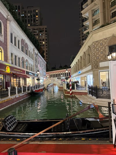 The image displays a nighttime scene within a commercial complex in Esenler, Turkey, designed with Venetian architectural elements around an artificial canal. The sky above is dark and overcast. Two rows of multi-story buildings flank a central waterway.

On the left, a building featuring light-colored stucco and red brick arches on its upper stories houses retail establishments at ground level, including "KAHVE DÜNYASI" and "ROMA". The building's roofline is adorned with dense green foliage. Behind this structure, a tall, modern high-rise building with numerous illuminated windows is visible against the dark sky.

On the right, another building with a patterned facade, arched windows, and decorative lampposts also features ground-level retail, including a store signed "KOTON". A group of approximately ten individuals is gathered on the illuminated walkway in front of this building. Behind this structure, another tall, modern high-rise building is partially visible.

The artificial canal, filled with dark green water, runs between these buildings. Its banks are lined with low brick walls and black metal railings. Reflections of the illuminated buildings and overhead lights shimmer on the water's surface. A white, arched pedestrian bridge spans the canal in the mid-ground. A red and white striped mooring pole stands on the right bank near the bridge.

Two black gondola-style boats are present. One is moored on the left side of the canal beneath the bridge, displaying a red interior. The second gondola is prominently featured in the immediate foreground, partially out of frame, showing a dark, tufted seat and a long wooden oar resting diagonally across its interior. This foreground boat appears to be positioned on or adjacent to a red carpeted surface. No individuals are visible in either gondola. The overall impression is that of a themed shopping and dining district.