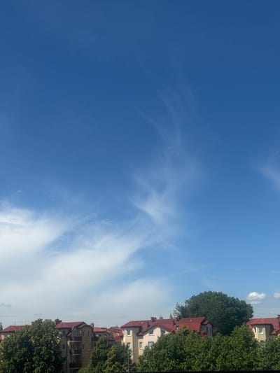 This is a media file showing a clear blue sky with wispy white clouds. On the right side of the image, several buildings with red roofs and light-colored facades are visible, interspersed with lush green trees. The scene appears to be residential. The lighting suggests it is daytime, likely mid-morning or afternoon, given the brightness of the sky and the shadows cast by the buildings. The weather is fair and sunny. The image is oriented vertically, with the sky occupying the left two-thirds and the buildings and trees the right third. There is no visible text in the image.