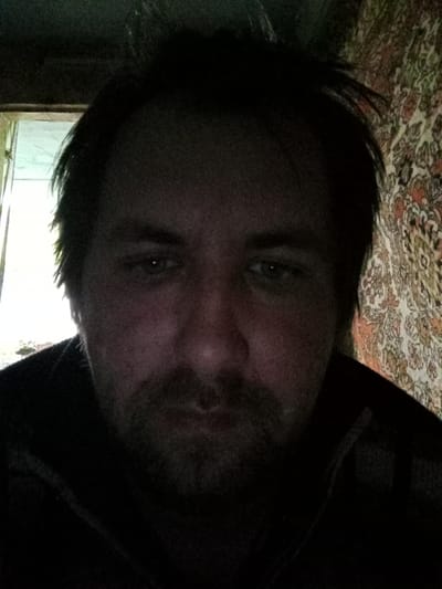 A dimly lit, close-up portrait captures a man with a beard and dark hair. His gaze is directed downwards. Behind him, a window provides a source of light. To the right, a patterned wall is visible. The location is assumed to be Parabel, Russia.