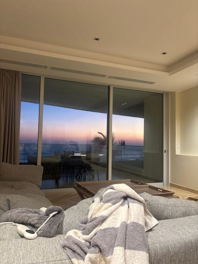 The image is taken indoors, likely from a high-rise apartment or hotel room, overlooking the ocean. The time appears to be during sunset or twilight, as indicated by the pink and orange hues in the sky and reflected in the ocean. A large window provides a view of the ocean and a palm tree silhouette is visible, suggesting a tropical location. In the foreground, a grey couch is partially visible with a blanket draped over it, along with a white charger plugged into what may be a heating pad. Outside the window, some kind of outdoor seating is visible with chairs and a table. Based on the presence of a tropical scene including the palm tree and ocean view, the location is likely Acapulco de Juárez, Mexico.