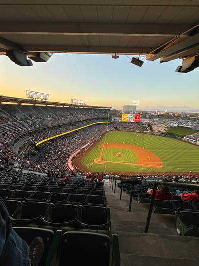 A baseball game is underway at Angel Stadium in Anaheim, California, United States of America. The sun is setting, casting a warm orange glow across the sky and illuminating the stadium. Spectators are seated in the stands, many wearing red, likely in support of the home team. The baseball field is green and well-lit, with players visible on the diamond. Beyond the stadium, buildings and distant mountains can be seen, indicating an urban environment. The perspective is from a high vantage point in the stands, looking down at the field and the surrounding cityscape.