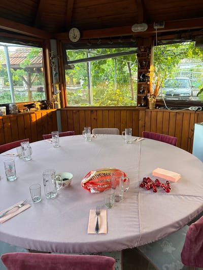 A round table is set with glasses, napkins, and cutlery for a meal. A pink tablecloth covers the table, and several red cherries and a small pink box are placed on top. A package of what appears to be crackers or cookies is also visible. The setting is an indoor area with wooden walls and large windows that look out onto a lush, green garden with grapevines. Sunlight streams through the windows, suggesting it's daytime. A vintage clock hangs on the wall. The overall atmosphere seems relaxed and possibly festive, as if preparing for a gathering or a meal. The crops show close-ups of the table settings and the outdoor view, highlighting the details of the food items and the natural surroundings. The image is taken from a slightly elevated angle, looking down at the table. There is text on the food package, but it is partially obscured and difficult to read clearly. The location is noted as Vaklino, Bulgaria.