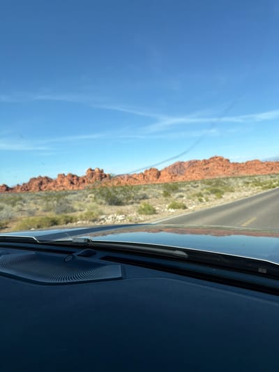 The image depicts a view through the windshield of a vehicle, likely a car. The exterior scene is of a landscape with red rock formations, sparse green vegetation, and a road. The sky is clear blue with some wispy clouds. The location appears to be in a desert environment, reminiscent of the southwestern United States. The road is asphalt, with a yellow line.
Inside the vehicle, part of the dashboard is visible. A black speaker is seen in the interior.
Unknown, United States of America is used as a stand-in for the specific location.