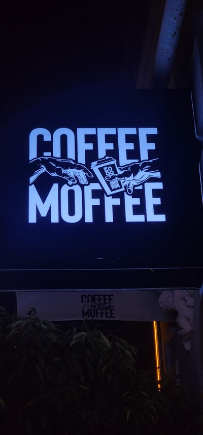 A neon sign for "COFFEE MOFFEE" is displayed on a dark, possibly outdoor, surface. The sign features white lettering and an illustration of two hands exchanging a coffee cup. Below the main sign, a smaller, darker sign with the same "COFFEE MOFFEE" logo is visible. This sign is partially obscured by lush green foliage in the foreground. The scene is lit by the neon sign and an orange vertical light source visible to the right, suggesting an evening or nighttime setting in an urban environment. The architectural elements visible, like a sculpted cherub figure on the right, suggest a place of business or establishment. The location is identified as Türkmenbaşy, Turkmenistan.