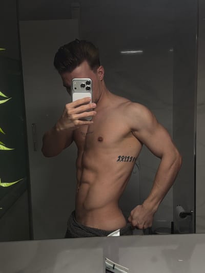 An adult male is captured in a mirror selfie within a modern bathroom, presumed to be located in Viseu, Portugal. The subject, who has a lean, muscular physique and short brown hair styled upwards, is bare-chested. He holds a dark grey towel around his lower torso with his right hand, which is visible gripping the towel's edge. He is taking the photograph with a silver-cased smartphone, equipped with a triple camera array, held by his left hand, which partially obscures his face. Distinctive body markings include a vertical chest tattoo on his left pectoral muscle, appearing to read "IMPERFECT" (reversed due to reflection), a horizontal tattoo of symbolic characters on his right rib cage, and a small barbell piercing on his right nipple. The environment, reflected in the mirror, features white tiled walls, a door handle or vertical fixture on the left, a toilet paper roll dispenser, and part of a toilet on the far right. A soft green reflection, possibly from foliage or a plant, is visible on the left side of the mirror.
