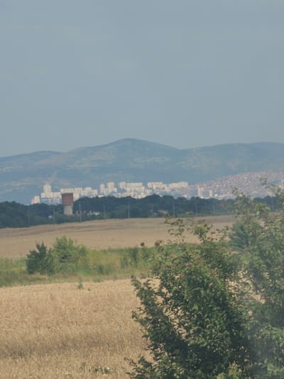 The image captures a rural landscape with a distant city skyline. In the foreground, a vast field of golden wheat stretches across the frame, with some scattered green vegetation and small white and blue wildflowers visible. A dense line of dark green trees borders the field, beyond which lies a water tower with a reddish-brown cylindrical structure atop a concrete base. Further in the distance, a sprawling city, identified as Bozduganovo, Bulgaria, is visible nestled against rolling hills. The city is characterized by numerous white, high-rise buildings and a dense collection of smaller structures, suggesting a significant urban area. The sky above is a hazy, pale blue, indicative of a clear but possibly humid day. The overall impression is one of a transition zone between rural agricultural land and a developed urban center, with the natural landscape giving way to man-made structures. There are no people or animals visible in the frame.
