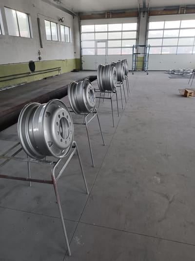 This media file presents an interior view of a large industrial workshop or garage located in Dobrodan, Bulgaria. The primary subjects are eight large, silver-grey metal wheels, likely for trucks or heavy vehicles, arranged in a line towards the center and right of the frame. Each wheel, appearing freshly painted or refurbished with a uniform matte metallic finish, rests on its own simple, slender metal stand, suggesting a stage in a drying, inspection, or processing operation.
The setting is an expansive, high-ceilinged industrial space. On the left, a concrete wall is painted white on top with a faded olive-green band along the lower section. Several large, rectangular windows punctuate this wall, allowing diffused natural light to illuminate the interior. Below the windows, a recessed area or pit, possibly for vehicle maintenance, is visible. On the right and rear of the image, two large, white sectional garage doors dominate, providing additional ample natural light and a soft, hazy view of the outside environment, hinting at trees or distant structures.
In the mid-ground, to the right of the row of wheels, a blue metal scaffolding unit on wheels is visible. Further into the background, stacks of round metal objects, possibly more wheels or components, are laid out on the floor, alongside a wooden pallet. The floor throughout the workshop is made of grey concrete, showing typical signs of wear, scuffs, and expansion joints.
The bright, yet soft and diffused, natural light streaming in from the windows and garage doors indicates that it is daytime. There are no people visible in the image, and no discernible text can be read on any objects or surfaces. The overall impression is one of an active, functional industrial environment focused on vehicle component maintenance or manufacturing.