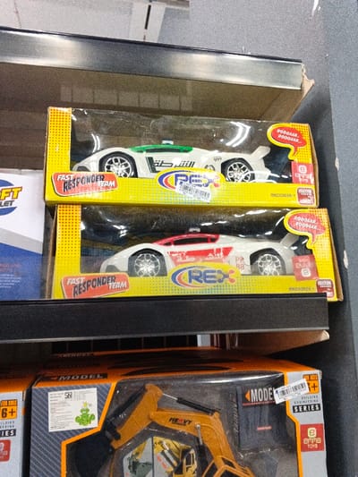 On a shelf, two toy cars are packaged in yellow boxes. Both are labeled "Fast Responder Team" and have "REX" logos. The top car is white with green accents and Arabic writing. The bottom car is white with red accents, also with Arabic writing. The boxes indicate they are "ROODAAR..." cars. Below them, a toy excavator is in an orange box, marked "Model Building Engineering Series" and suitable for ages 6+.