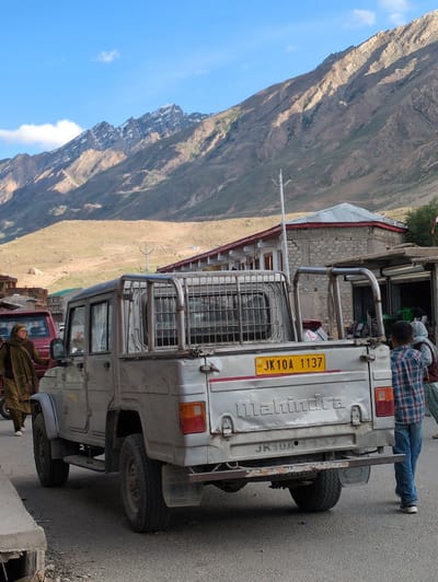 In Padum, India, a silver Mahindra pickup truck is parked on an asphalt road. The truck's rear is visible, displaying a yellow license plate that reads "JK 10A 1137". The word "Mahindra" is embossed on the tailgate. To the left of the truck, a woman wearing a green shawl and a brown outfit is partially visible, standing near a red vehicle. To the right of the truck, a young boy in a plaid shirt and blue jeans walks away from the camera. Behind the truck, buildings with corrugated metal roofs and stone walls are present, nestled against a backdrop of rugged, sun-drenched mountains with patches of snow on their peaks. The sky above is a clear blue with a few wispy clouds. The scene appears to be a street in a town or village, with the natural landscape dominating the background.