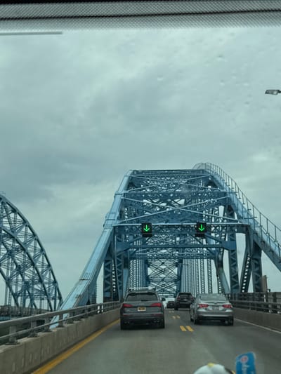 The image shows a view from inside a vehicle traveling on a bridge, likely during the day under overcast skies. The bridge is a large steel truss structure, painted blue, and appears to be quite old. Traffic is visible on the bridge, with several cars driving ahead. Green arrow traffic lights are visible, indicating the direction of traffic flow. A portion of the vehicle's windshield wiper or frame is visible on the left side of the image. The sky is a uniform gray, suggesting cloudy weather, possibly with rain or fog. The road surface is asphalt with yellow lane markings. There is no visible text in the image. The scene evokes a sense of travel and transit, with the imposing bridge structure and the steady flow of vehicles. The location is identified as the Town of Tonawanda, United States of America.