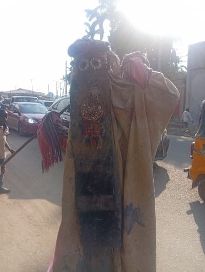 The image depicts a street scene at Itakpe Junction, Nigeria, centered on a tall, upright costumed figure. This figure is draped in voluminous, light brown fabric, possibly sackcloth, showing signs of wear and dark stains. Its headpiece is dark and textured, featuring two distinct white, circular, floral-like embellishments. A long, dark vertical panel adorns the front of the figure, embellished with metallic, circular objects resembling coins, and woven red and dark patterns. A fringed, striped fabric piece hangs from the figure's left side. To the immediate left of the costumed figure, a person, likely a young male wearing a light shirt and dark trousers, holds a long, dark stick. In the left background, several vehicles, including a red sedan, are visible on a paved road, indicating traffic. On the right, the rear portion of a yellow motorized tricycle (Keke Napep) is partially visible on the road. A smaller figure walks in the distant right background near a light-colored building. The sky is bright with prominent sun glare originating from the upper right, and palm trees are visible against the sky. The ground comprises both light-colored dirt and paved asphalt.