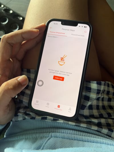 A person's hand, with short fingernails, is holding a black-rimmed smartphone. The phone displays the ShopeeFood app, specifically the "Pesan Saya" (My Orders) screen. The screen shows a "Connection Failed" message in Indonesian: "Koneksi gagal. Mohon periksa jaringanmu dan coba lagi." (Connection failed. Please check your network and try again.) Below this message is a red button labeled "Coba Lagi" (Try Again). The phone is resting on a person's lap, who is wearing light blue sweatpants and a dark blue and white checkered fabric. The background is out of focus, showing a blurred texture. The image appears to be taken indoors, possibly in a home or public space. There are no explicit indicators to pinpoint the location to Pekalongan, Indonesia, or any specific city.