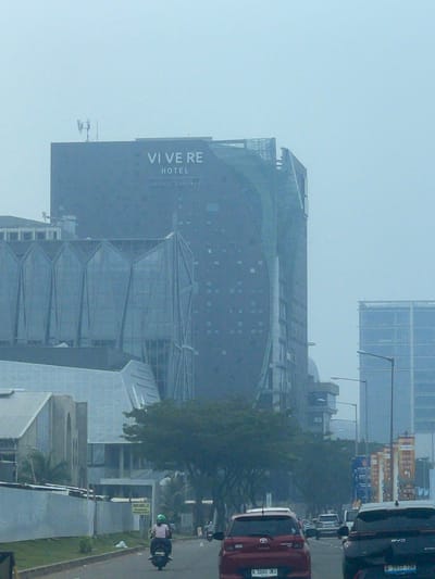The image shows a street scene in the city of Medan, Indonesia, likely on a hazy or overcast day. In the background, a large, modern, dark-colored building with "VI VE RE HOTEL" prominently displayed on its facade dominates the view. To the left, a building with a distinctive geometric glass facade is visible. Further to the right, another tall, glass-enclosed building stands. In the foreground, a paved road is lined with trees and other vegetation. Several vehicles are present on the road, including a red car, a black SUV, and a motorcycle with a rider wearing a green helmet and pink shirt. Signs are visible along the roadside, including one that appears to say "HATI-HATI," which means "Be careful" in Indonesian. The overall atmosphere is urban, with a sense of ongoing activity and development.