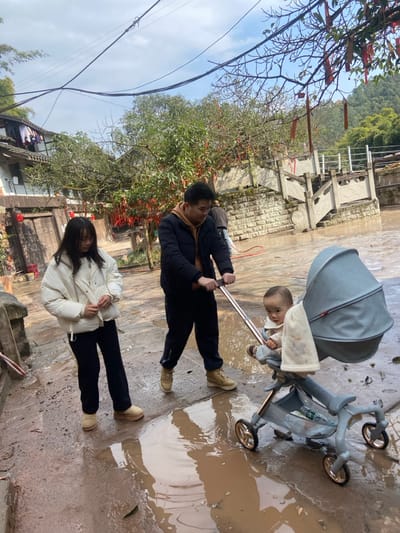 A family of three on a paved surface with puddles is visible. A man in a black jacket pushes a stroller containing a baby. A woman wearing a white jacket stands near the stroller. Trees are in the background along with buildings. Overhead power lines are visible, as well as trees with decorative red strips hanging from them.