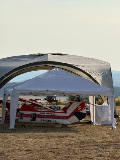 An outdoor scene depicts a large, model helicopter with "COLUMBIA" and "P2-CHE" markings, positioned under two tents. The helicopter is white and red, with visible windows and a rotor. The tents are white; one is a pop-up canopy, and the other is a larger, arched structure. The ground is covered in dry, brown grass. In the background, rolling hills and a clear sky are visible, with a distant wind turbine on the right. No people are present in the immediate vicinity of the helicopter. The overall impression is one of an organized outdoor event or display, possibly related to model aviation.