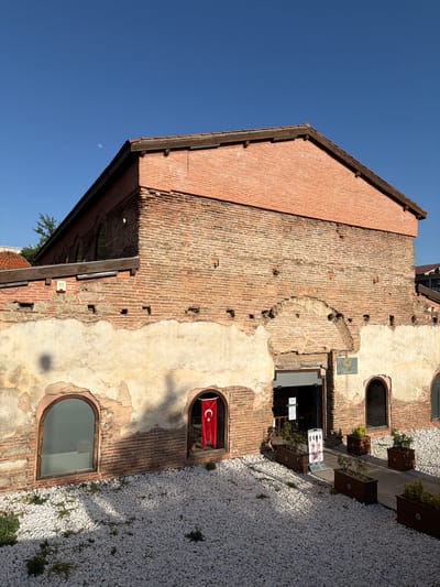 The image displays an old, brick building with a rustic and weathered facade under a clear blue sky. The building, likely several stories high, features a textured exterior where sections of exposed reddish-brown brickwork alternate with patches of light-colored, crumbling plaster. Its roofline, visible at the top, is made of reddish tiles and dark wooden trim.

On the building's right side, several arched windows and an open doorway are visible. A vibrant red Turkish flag, featuring a white star and crescent, hangs prominently in one of the arched windows, serving as a clear national identifier. Below this, another arched window reflects the surroundings. A larger, open doorway leads into the building, hinting at an interior space, possibly a commercial establishment. Next to this entrance, a white sign displays multiple international "no" symbols, including prohibitions against smoking, food/drink, and pets. Further up the wall, a small, square, greenish plaque is affixed, and below the doorway, a small white sign with text is present, though the text is not clearly legible.

To the immediate right of the building, a light-colored ground cover of small, rounded white gravel extends, interspersed with sparse green vegetation. This area appears to be a pathway or decorative courtyard. A wooden deck or pathway runs parallel to the building, featuring several dark wooden planters holding small green plants.

The sky, a brilliant and clear blue, dominates the upper portion of the scene. Faintly visible in the upper right quadrant of the sky is a crescent moon, suggesting a daytime shot. The bright sunlight casts distinct shadows, indicating a sunny day, likely mid-morning or afternoon. The overall atmosphere is quiet and still, emphasizing the historical character of the architecture in Iznik, Turkey.