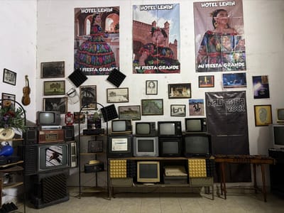 An eccentric interior showcases a collection of vintage electronics. Old television sets are stacked on shelving units, interspersed with audio equipment and various framed pictures adorning the wall behind. Posters advertising "Hotel Lenin" with images of women in traditional dresses are prominent above. Additional wall decor includes black sound-absorbing panels, and a black banner with "Naupaca Rock." A violin hangs on the left wall, and a xylophone stands on the right, adding to the room's eclectic character.