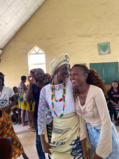 The image depicts a gathering of people, primarily women, in an indoor setting that appears to be a community hall or church in Dutsen Kato, Nigeria. The room has pale yellow walls and a tiled floor. A small, diamond-shaped window with a light-colored frame is visible, suggesting natural light entering the space. There is a green window with multiple panes above a person seated in the background. On the wall, a square emblem with a cross and some text is displayed.
Several women are present, some facing the camera and others turned away. The woman in the foreground is leaning in to speak to an older woman, and both are smiling, conveying a sense of warmth and connection. The older woman is wearing a headscarf and a white lacy top, adorned with a colorful beaded necklace featuring red, yellow, green, and black beads. The younger woman is wearing a light-colored long-sleeved shirt with a butterfly design on it and jeans. Other women in the background are dressed in vibrant, patterned African attire. The overall mood seems joyful and social, possibly a celebration or a community meeting.
The image is oriented vertically, suggesting it might have been taken in portrait mode or that the camera was held at an angle. The lighting is somewhat uneven, with some areas brighter than others. The time of day appears to be daytime due to the natural light from the window. Visible text includes "VICTORIA" on a black t-shirt worn by a person in the background, and partial text on the beaded necklace and on the emblem on the wall, which is difficult to decipher clearly. The presence of traditional clothing and the general atmosphere suggest a cultural or religious event.