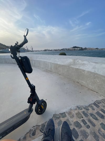 An electric scooter is parked on a paved walkway next to a body of water. The walkway transitions from concrete to cobblestones. In the background, across the water, a city skyline is visible with buildings and hills. The sky is partly cloudy. A person's feet, wearing dark athletic shoes, are visible at the bottom of the frame. The scene appears to be in Cartagena, Spain, likely along the waterfront.