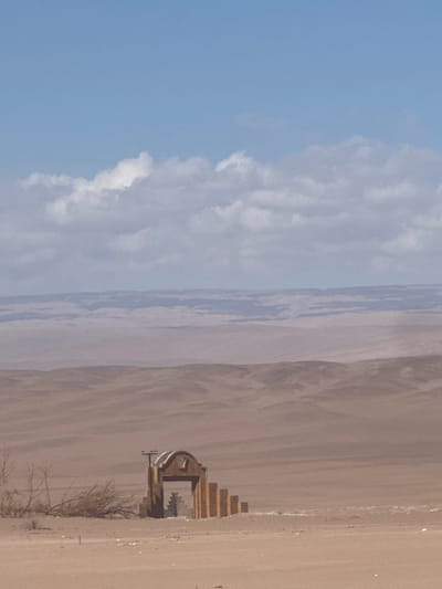 The image displays an expansive, arid desert landscape, characteristic of the region near Pica, Chile, under a mostly clear blue sky with a band of scattered white and grey clouds across the mid-upper portion. In the immediate foreground, a weathered, light-colored architectural structure, resembling a gate or archway, stands partially submerged in the sand. This structure exhibits an arched opening, a decorative, stylized plant or starburst-like pattern within the arch, and a stepped profile on its right side. A dark, horizontal bar is mounted atop the structure. To the left of the structure, several dried, leafless branches and sparse vegetation are visible, along with minor debris on the sandy ground. The midground and background are dominated by successive ranges of rolling desert hills and plains, receding into a hazy distance. No individuals, animals, or active subjects are present in the scene; it is static and devoid of overt human activity.