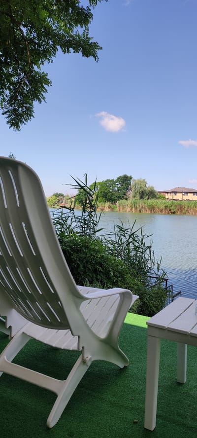 A white plastic lounge chair is in the foreground, angled to the left and partially cropped. The chair is situated on a green artificial turf surface. To the right of the chair, a portion of a white wooden table is visible. Beyond the chair and table, a body of water, likely a river or canal, stretches across the middle ground. The water is calm with a slight ripple. A dense growth of green reeds and other vegetation lines the opposite bank of the water. Several trees with lush green foliage are present behind the reeds, and in the distance, two houses with beige and yellow siding are visible. The sky above is a clear, bright blue with a few small, wispy white clouds. A portion of a leafy green tree branch extends from the upper left corner of the frame. The scene appears to be an outdoor recreational area, possibly a resort or park, in the city of Dinskaya, Russia.