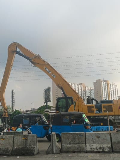 A daytime, medium-shot photograph captures an urban scene in the Special Capital Region of Jakarta, Indonesia, featuring heavy machinery, local transport, and residential infrastructure under an overcast sky. In the foreground, a series of grey concrete Jersey barriers demarcate an area. Behind these barriers, two light blue, three-wheeled Bajaj vehicles are positioned, suggesting their use as local auto-rickshaws. A single individual, wearing a light blue top, is visible inside the left Bajaj, facing left.
Dominating the central and right areas of the image is a large yellow "POWERPLUS" brand excavator, model "PP 600E-XI," distinguished by its extended, articulated arm angled upwards and to the left. The midground features dense green foliage, several vertical utility or tower structures, and a prominent billboard frame. An elevated road structure is partially visible beyond these elements. The background is composed of multiple tall, light-colored apartment or residential buildings, indicative of high-density urban housing. Numerous horizontal utility lines or power cables stretch across the upper portion of the frame. The sky is uniformly light grey and overcast, providing diffuse ambient lighting. The scene appears static, with no overt actions or interactions depicted.