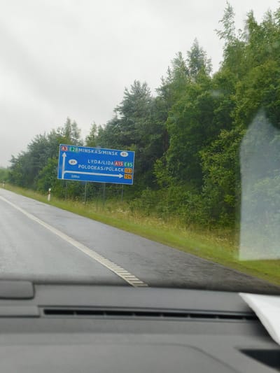 An eye-level, medium shot captures a blue road sign on the right side of a two-lane asphalt road. The sky is overcast, and trees line the right side of the road. The sign has white text and arrows indicating directions. The top portion of the sign reads "A3 E28 MINSKAS/MINSK BY" with an arrow pointing straight ahead. Below that, it reads "BY LYDA/LIDA A15 E85" with an arrow pointing to the right. Further down, it states "POLOCKAS/POLACK 103 106" with an arrow pointing to the right. A smaller section of the sign reads "500m" with an arrow pointing straight ahead. The road is wet, suggesting recent rain. The lower portion of the image shows the dashboard of a vehicle. The scene is set in Skaidiškės, Lithuania.