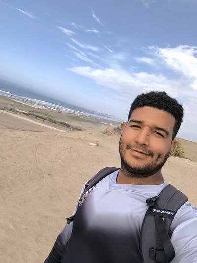 A medium-shot selfie of a man with dark hair and a beard, wearing a grey and black shirt and a black backpack, smiling at the camera. The background features a sandy dune landscape, a view of the ocean, a coastal cityscape, and a partly cloudy sky. The city is likely Huanchaco, Peru, given the characteristics of the terrain and coastal development visible.