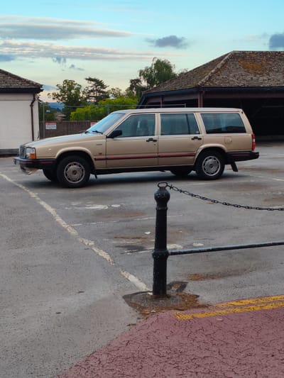 The image depicts a beige Volvo 700 series station wagon parked in an outdoor paved area. The car, viewed from its left side, is positioned in what appears to be a parking lot or an open yard. In the background, there are buildings with tiled roofs and some trees. The sky is visible with scattered clouds. A black metal bollard with a chain is in the foreground, separating the parking area from a textured red surface with yellow lines. The scene is set during the day, likely in the United Kingdom, given the context of Wychavon. No people are present in the frame.